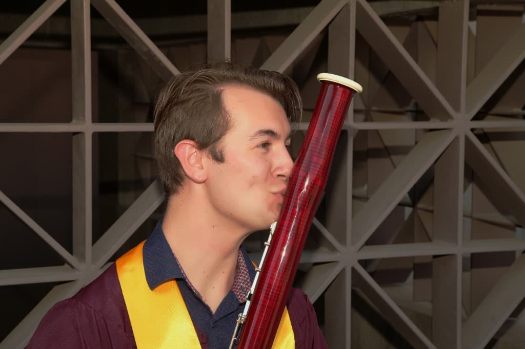 Mason Cox in ASU maroon and gold graduation regalia, playfully posing with his bassoon