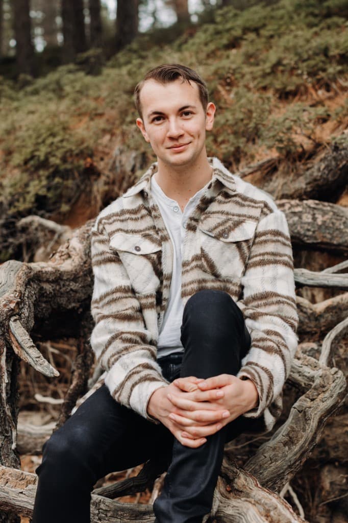 Mason Alexander Cox, bassoonist and composer, seated outdoors on tree roots in a wooded setting